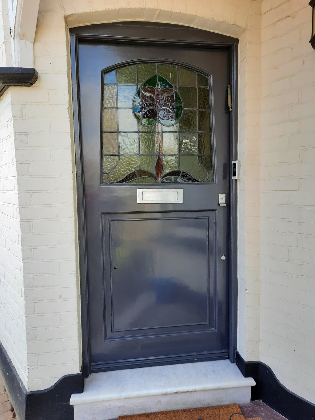 A dark charcoal gray front door with a decorative stained glass window featuring a bird motif, set in a white brick wall with a small stone step.